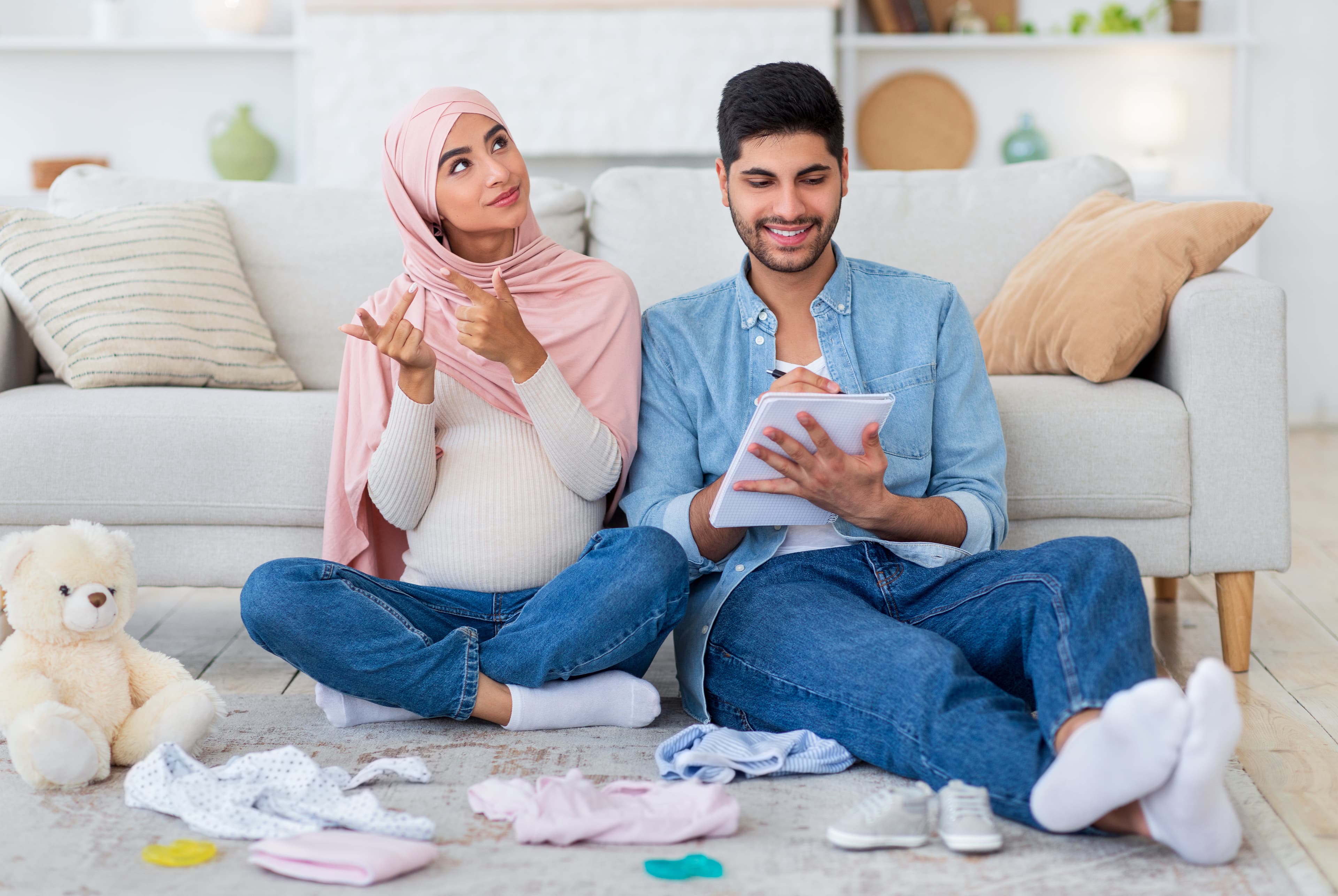An expectant couple sitting on the ground next to a pile of baby clothes.