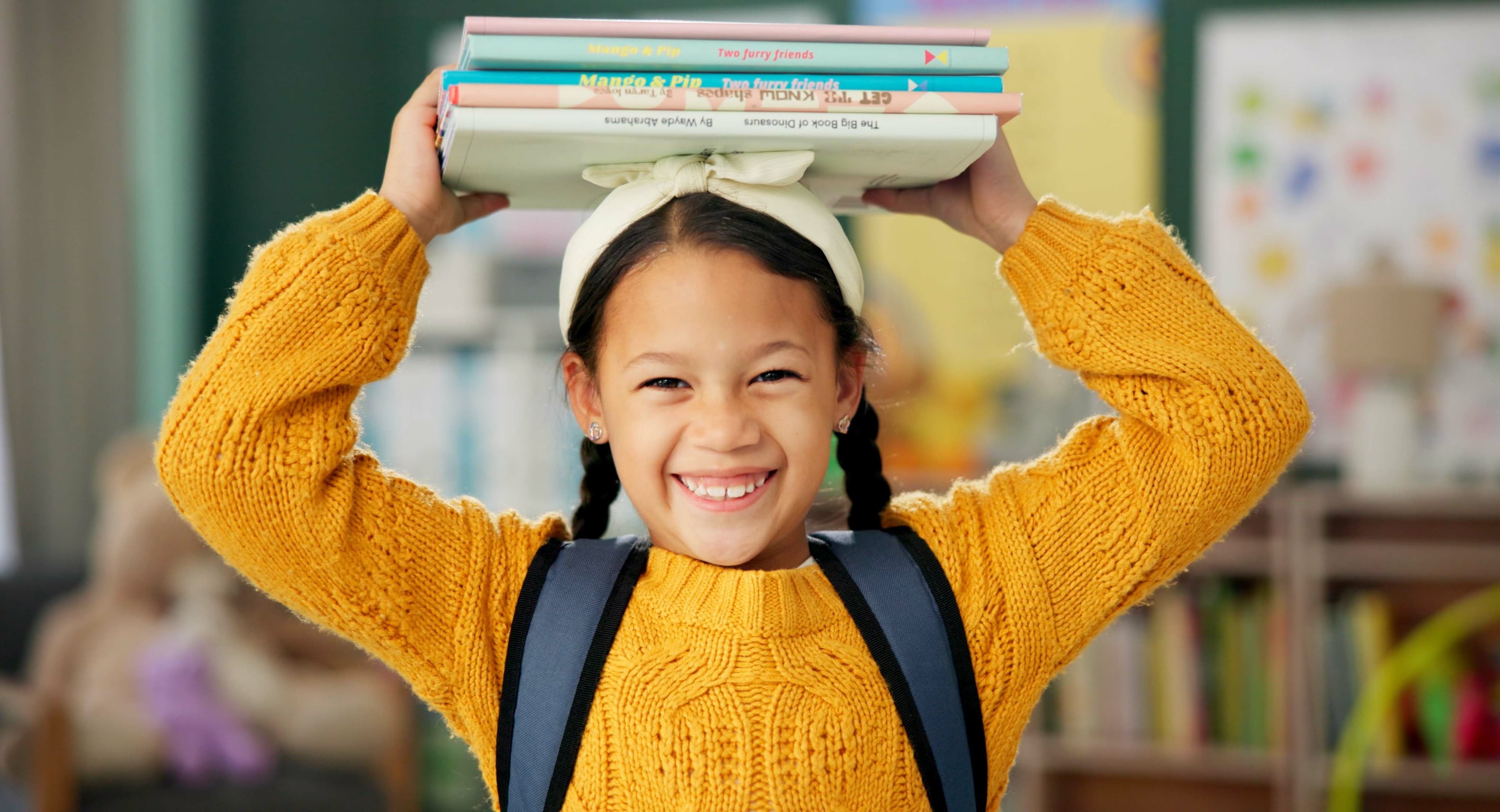 A smiling girl with a yellow shirt carrying books on top of her head