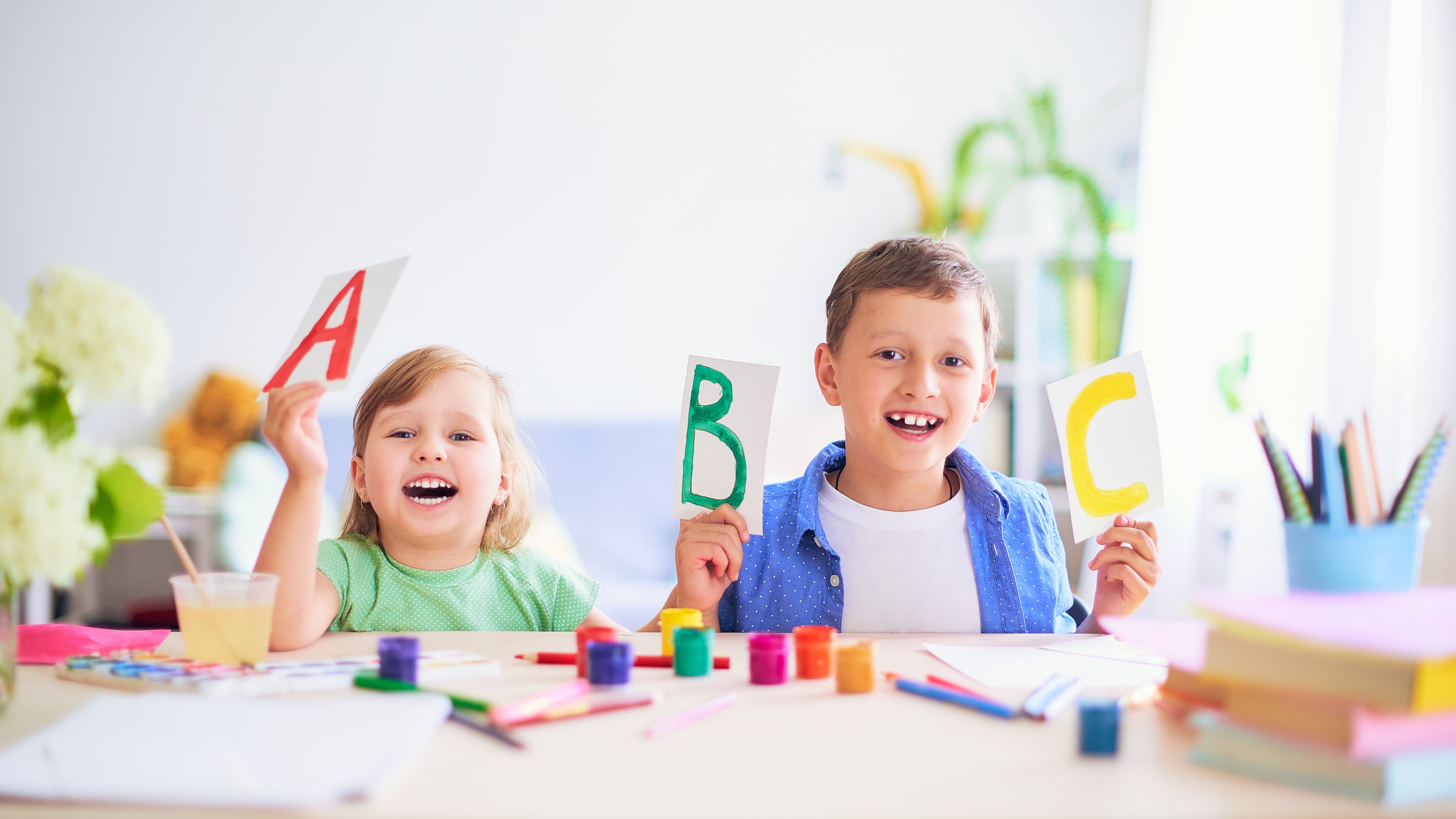 Two smiling children painting at a table holding up their paintings.